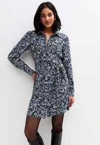 Ung kvinna med mörkt hår iklädd en långärmad svart klänning med små blå och vita blommor, svarta strumpbyxor och svarta knähöga stövlar.