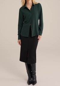 Zielona koszula na guziki z długimi rękawami, zestawiona z dopasowaną czarną spódnicą z przednim rozcięciem oraz czarnymi, sięgającymi kolan, butami na obcasie.