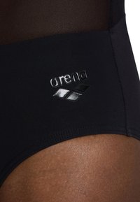 Crni donji dio kupaće gaće s glatkom tkaninom, mrežastim panelima na vrhu i istaknutim, sjajnim logotipom s dizajnom vala sa strane.