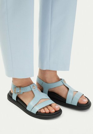 Hellblaue Ledersandalen mit schwarzen Sohlen und verstellbaren Schnallenriemen, getragen zu hellblauen Hosen an nackten Füßen.