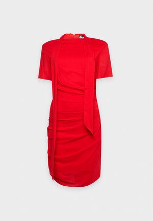 Une femme porte une robe rouge drapée avec un long lien noué autour du cou. Elle tient un sac à main marron foncé et porte des escarpins noirs à brides.