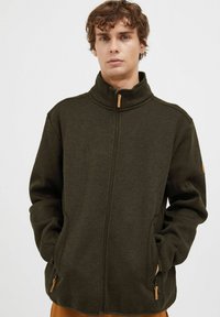 Tamnozeleni zip-up jakna s visokim ovratnikom, bočnim džepovima i teksturiranim materijalom. Ima drveni zip pull i suptilne detalje u šivanju.
