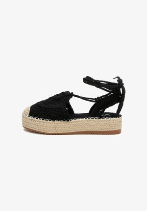 Espadrille negro tejido con punta redonda, suela intermedia de yute natural y lazos negros. Textura detallada en la parte superior y acentos decorativos.