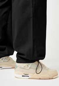 Beige Sneakers mit Mesh-Akzenten und sichtbarer Luftpolsterung, kombiniert mit schwarzen, locker sitzenden Hosen. Die Sneakers sind mit "AIR" Branding versehen.