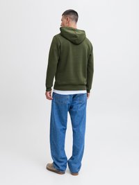 Olivovo zelená mikina s kapucňou s jemnou textúrou, v kombinácii s voľne strihanými modrými džínsami, ktoré majú zadné vrecká a svetlú pranie.