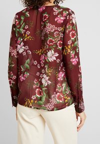 Kobieta w długim, przezroczystym bordowym bluzce z różowo-białym kwiatowym wzorem, w połączeniu z beżowymi spodniami, widoczna z tyłu.