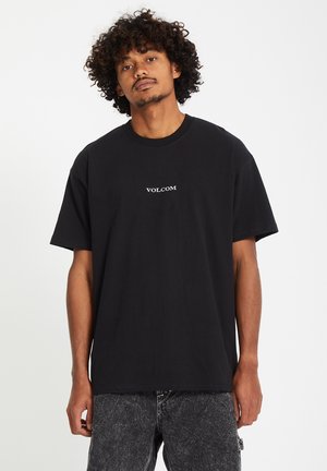 Zwart katoenen t-shirt met ronde hals, korte mouwen en een wit "VOLCOM" logo op de borst. Gekleed met lichtgrijze versleten jeans.