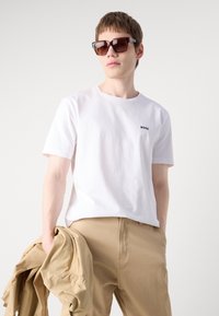 Tricou alb din bumbac cu logo-ul "BOSS" pe piept, asortat cu pantaloni bej și ochelari de soare pătrați maroon, ținut cu o jachetă bej.