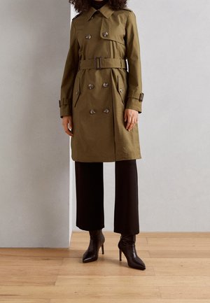 Persona con gabardina verde oliva con cinturón, pantalones negros de pierna ancha y botas negras de tacón con punta afilada, de pie sobre suelo de madera.