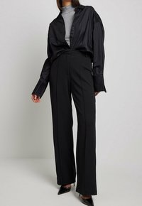 Schwarzes, übergroßes Hemd mit Knöpfen, kombiniert mit hoch taillierten, weiten schwarzen Hosen. Das Modell trägt spitze schwarze Absätze und darunter einen strukturierten grauen Rollkragenpullover.