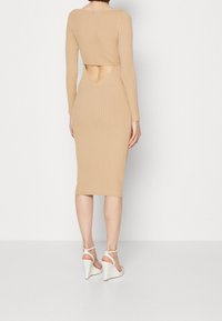 Robe midi beige côtelée avec des manches longues, détail dos nu et silhouette ajustée. Des sandales compensées avec des brides aux chevilles sont portées.