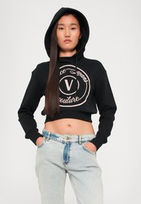 Juodas trumpas hoodie su dideliu apvaliu logotipu, kuriame yra "V" smėlio spalvos. Pora su šviesiai mėlynomis džinsinėmis kelnėmis, turinčiomis išblukusį dizainą.
