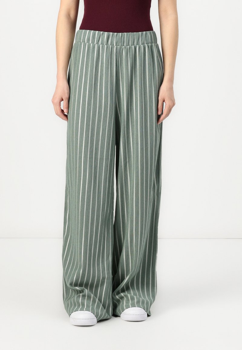 Femme portant un pantalon large à rayures verticales vertes et blanches, taille élastique, et des baskets blanches, debout sur un fond blanc.