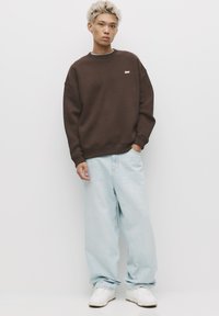 Rjava bombažna jopa z okroglo izrezom, sproščenim krojem in majhnim vezenim logotipom. Kombinirano s svetlo modrimi širokimi kavbojkami in belimi supergami.