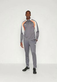 Homem em pé, vestindo um fato de treino cinzento da Puma com riscas laranja e branca, e sapatilhas pretas de desporto, em frente a um fundo simples.