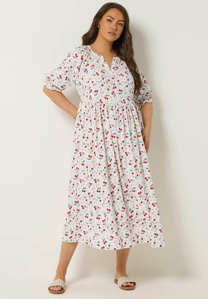 Kobieta w białej sukience midi z różowo-czerwonym nadrukiem wiśni, z bufkami na rękawach i klapkami na nogach, stojąca na tle jednolitego jasnego tła.