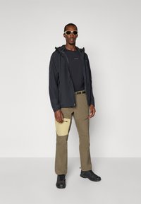 Schwarze wasserdichte Jacke über einem dunklen Shirt, olivgrüne Cargo-Hosen mit einer hellgelben Tasche, schwarze Schuhe und Sonnenbrille, stehend vor einem weißen Hintergrund.