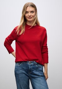 Maglione rosso a maglia con polsini a coste e scollatura a girocollo, abbinato a jeans in denim blu con tasca frontale e leggera scoloritura.