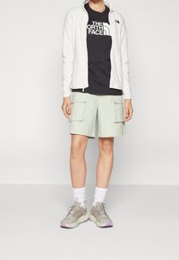 Giacca bianca con zip, t-shirt nera con logo, pantaloni cargo verde chiaro, calzini bianchi e scarpe da ginnastica grigie con dettagli viola.