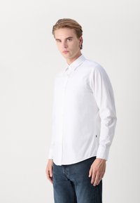 Camisa blanca de manga larga con cuello abotonado, que presenta un corte entallado y un ligero brillo. Combinada con jeans de corte oscuro, de textura suave.