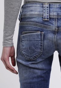 Lähikuva kädestä sinisen farkkukankaan takataskun vieressä, jossa on ompeluyksityiskohtia. Kädessä on päällä harmaa ribatoppi pitkällä hihalla.