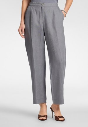 Vrouw draagt grijze hoge taille, recht gesneden broek met knoopsluiting, gecombineerd met bruine open teen hoge hak sandalen op een effen achtergrond.