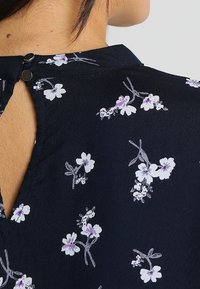 Blouse marine avec un imprimé floral comprenant des fleurs blanches et violet clair. Dos à découpes avec deux boutons noirs. Texture légère et lisse.