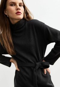 Zwarte coltrui jurk in een gestructureerde stof, met een ceintuur in de taille en lange mouwen. Het ontwerp heeft een losse pasvorm en subtiele ribbels.