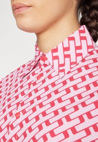 Button-up shirt in pink with a bold red geometric pattern. Textured fabric with a classic collar and rounded hemline. Red buttons feature.