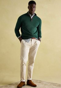 Zaļš zipa kakla džemperis virs zila un balta pārbaudes krekla, krēmkrāsas krokodila auduma bikses un brūni zamšādas potīšu zābaki. Strukturēta piegriezuma ar ribotām aprocēm.