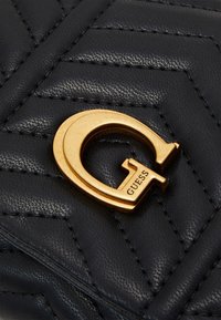 Black quilted leather material with a prominent gold circular logo featuring the brand name "GUESS" embossed. Textured surface detail.