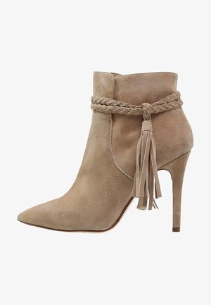 Bottine en daim beige avec un bout pointu, des détails tressés autour de la cheville et un accent de pompon, reposant sur un talon aiguille.