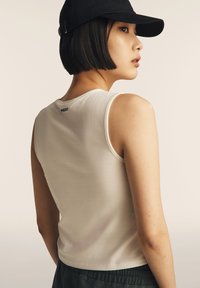 Giovane donna con capelli neri corti, che indossa un cappellino nero, una canotta bianca senza maniche e pantaloni scuri, di profilo contro uno sfondo neutro.