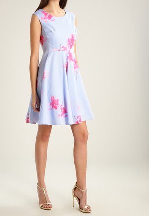 Kobieta ubrana w bezrękawnikową, jasnoniebieską sukienkę w różowe wzory kwiatowe, w połączeniu z beżowymi sandałami na wysokim obcasie, stojąca na jednolitym tle.