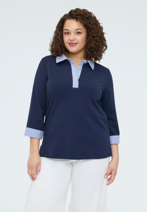 Žena s kudrnatými vlasy, oblečená v tmavě modrém polokošili s dlouhým rukávem, pruhovaným límečkem a manžetami, a bílých kalhotách, stojící před jednobarevným pozadím.