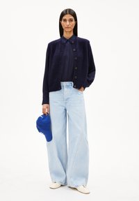 Marineblauer Strickweste mit Knöpfen über einem dunkelblauen Hemd, kombiniert mit hellblauen, weit geschnittenen Jeans und einer blauen Mütze, dazu beige Schuhe.