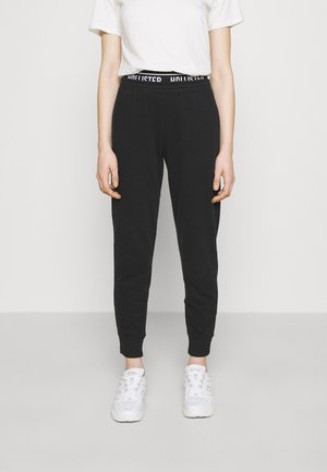 Persoon draagt zwarte joggingbroek met zichtbare Hollister tailleband, wit T-shirt en witte sneakers, staand tegen een effen achtergrond.