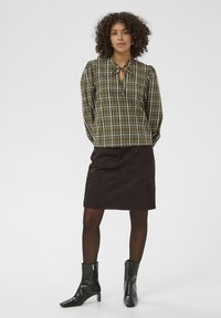 Chemise à carreaux verts avec un col en forme de cravate, associée à une jupe en daim marron et des bottines noires. Des collants foncés complètent le look.