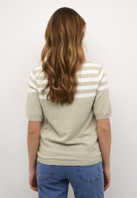 Kvinde med langt bølget brunt hår iført en beige og hvid stribet kortærmet bluse og blå jeans, stående foran en ensfarvet baggrund.