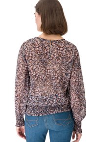 Femme portant un chemisier à manches longues à motif floral avec des poignets et un ourlet froncés, associé à un jean bleu, vue de dos.