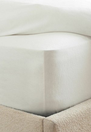 Nahaufnahme einer Bettecke mit weißem Spannbettlaken und einem strukturierten beigen gepolsterten Bettrahmen.