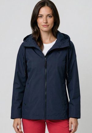 Frau trägt eine marineblaue Kapuzenjacke über einem weißen Hemd und roten Hosen, schaut nach vorne mit neutralem Ausdruck.