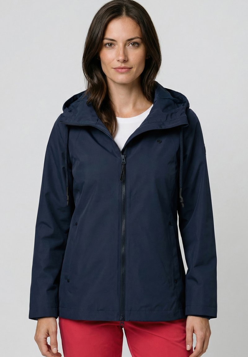 Vrouw draagt een marineblauwe jas met capuchon over een wit shirt en rode broek, kijkt recht vooruit met een neutrale uitdrukking.