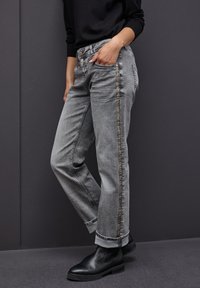 Vaqueros de mezclilla gris con corte recto, que presentan una franja decorativa de cuentas a lo largo del costado y puños enrollados, combinados con botas negras hasta el tobillo.
