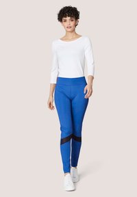 Blauwe actieve leggings met een gaasaccent bij de onderbenen, gecombineerd met een witte lange mouwtop en witte sneakers. Gladde textuur, rekbaar stof.