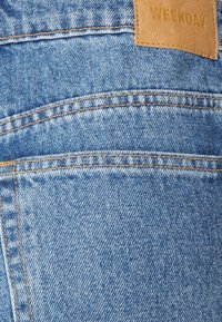Tecido de denim azul com textura e padrões visíveis; apresenta costuras amarelas e uma etiqueta de couro castanho em relevo com a inscrição "WEEKDAY."