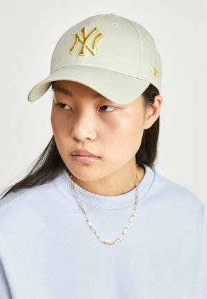 Giovane con cappellino beige con logo dorato di New York, felpa azzurro chiaro e collana dorata, che guarda leggermente di lato.