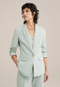 Světle zelený blazer s přizpůsobeným střihem, klopovými límci a dvěma předními kapsami. Disponuje jedním knoflíkem na zapínání a ohrnutými rukávy.