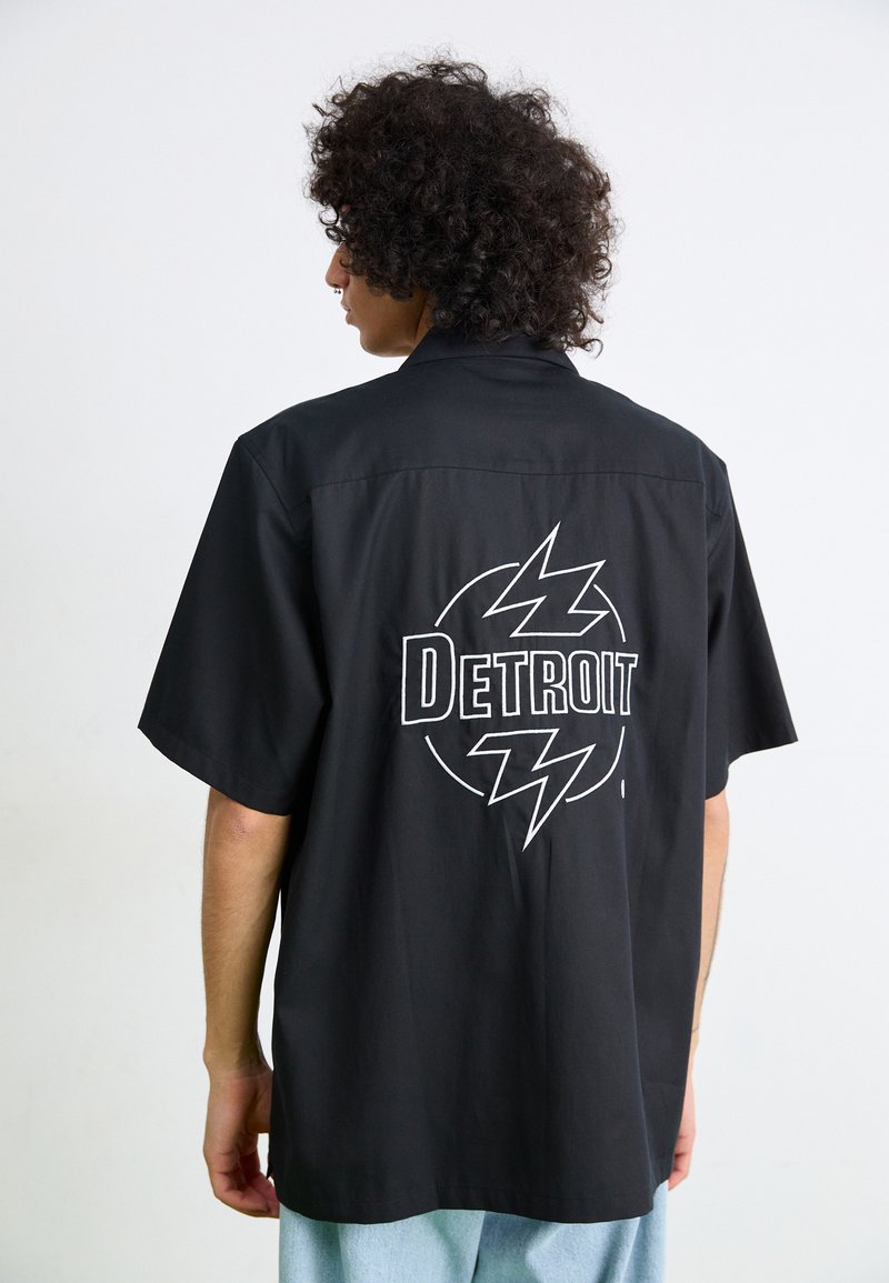 Svart kortermet skjorte med en rygglogo som leser "DETROIT" og lynmotiver, laget av glatt stoff med en avslappet passform.