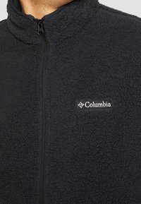 Giacca in pile nera con colletto alto, dotata di zip completa e di un piccolo logo Columbia sul lato sinistro del petto. Tessuto morbido e texture.